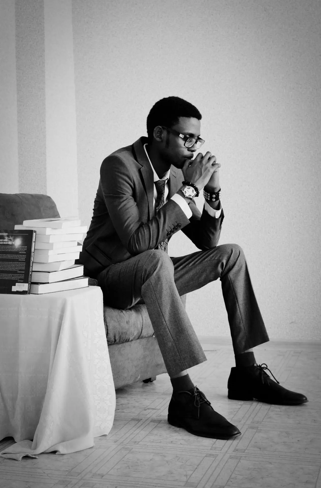 A thoughtful man in formal wear sits indoors, surrounded by books, conveying a pensive mood.